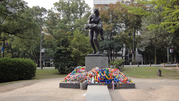 chains of paper cranes piled at the foot of a statue of a woman carrying a child